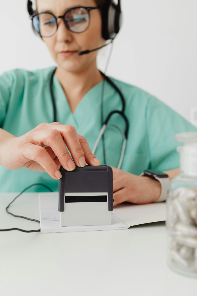 Female healthcare worker in scrubs using an office stamp, wearing headphones and eyeglasses.