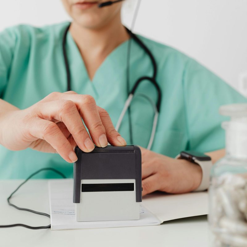 Female healthcare worker in scrubs using an office stamp, wearing headphones and eyeglasses.